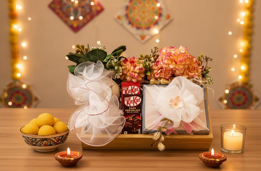 Gift basket with flowers, chocolates, and a candle on a wooden table with decorative lights in the background.