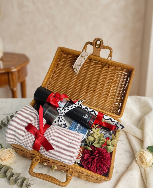 Wicker picnic basket with items including a bottle, wrapped gifts, and flowers on a table.